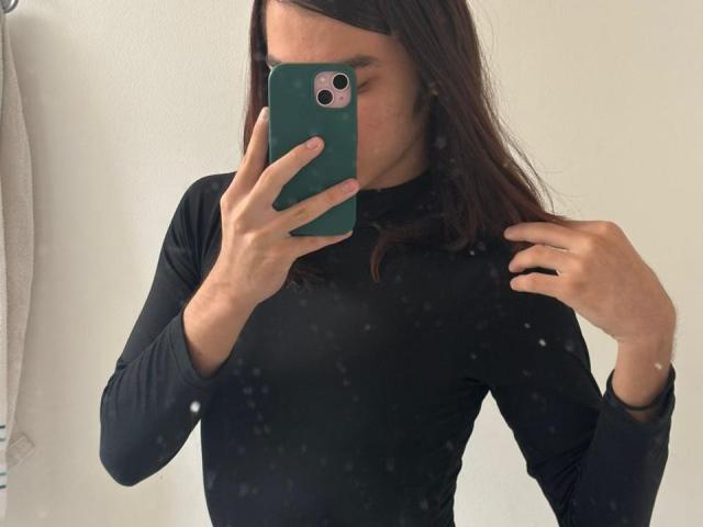 TamaraGarcia with medium brown hair, wearing a tight black long-sleeve top, taking a mirror selfie with a green phone, hands near her face, showing small tits.