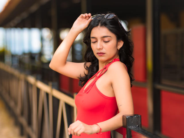 JazminSweet69 with wavy black hair, wearing a red halter top, adjusting sunglasses on her head, standing by a black railing, looking down with a calm expression.