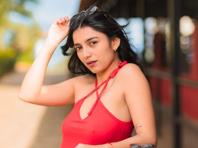 JazminSweet69 with medium tits in a red halter top, black hair, brown eyes, and a small tattoo on her right arm, holding sunglasses above her head.