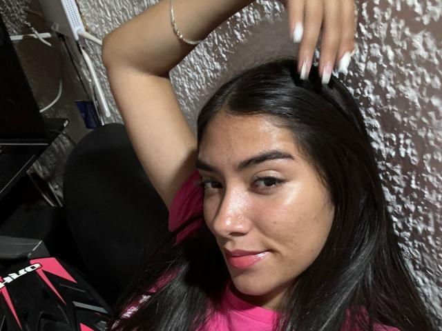 SharonKlart with long black hair, pink shirt, shiny face, arm raised, white nails, bracelet, slightly smiling, sitting in black chair, right hand touching head.