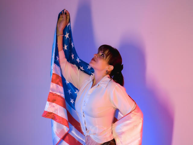 MilenaHott69 with her tits up, holding an American flag, wearing a white button-up shirt, looking up, shadow on wall, pink and blue lighting, hands on hips.