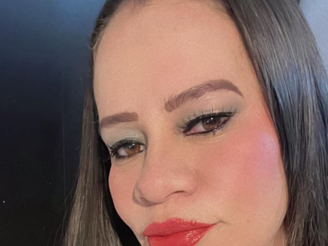 CharllotI's face, showing brown eyes with green eyeshadow, red lipstick, and slightly flushed cheeks. She has dark brown hair, thick eyebrows, and a subtle smile.
