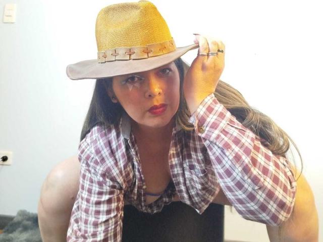 ZafiroYeux indossa un cappello di paglia beige con una fascia a motivi a croce, una camicia a quadri e rossetto rosso, è piegata in avanti con lunghi capelli castani, con il décolleté visibile e un anello alla mano destra..