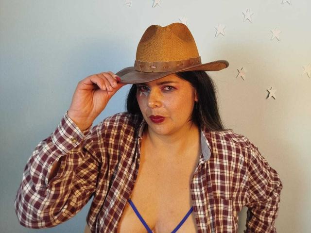 ZafiroYeux wearing a brown hat, plaid shirt, and blue bra, showing her tits, with dark hair, red lipstick, and a confident expression, touching her hat.