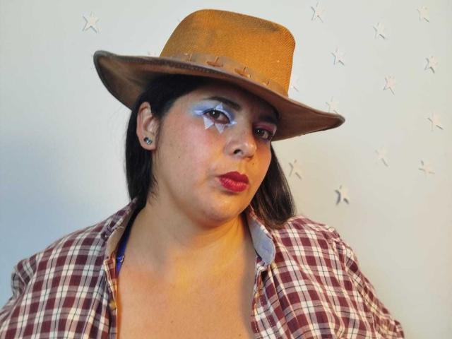 ZafiroYeux con un cappello marrone, trucco bianco con stelline sugli occhi, rossetto rosso, camicia a quadri e piccoli orecchini, che guarda leggermente di lato con un'espressione seria..