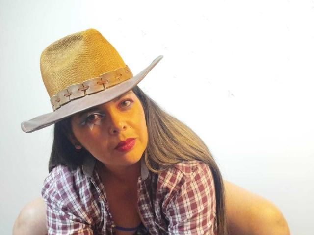 ZafiroYeux with tan skin, red lipstick, and long brown hair, wearing a brown straw hat with cross-stitched band, and a red and white plaid shirt, looking sideways.