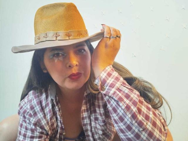 ZafiroYeux with brown wavy hair, wearing a tan straw hat with a woven band, red plaid shirt, and red lipstick, touching hat with right hand, showing a ring.