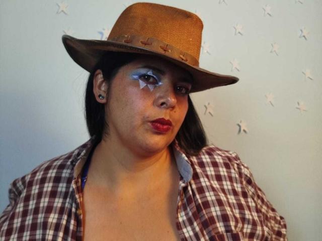 ZafiroYeux has dark hair, a brown hat with star decorations, blue star makeup on her cheeks, red lipstick, and a brown and white plaid shirt.