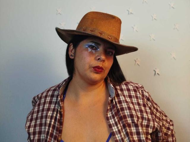 ZafiroYeux con cappello marrone, trucco bianco sul viso, rossetto rosso, camicia a quadri, reggiseno blu e orecchini, che mostra un'espressione seria, con il petto scoperto..
