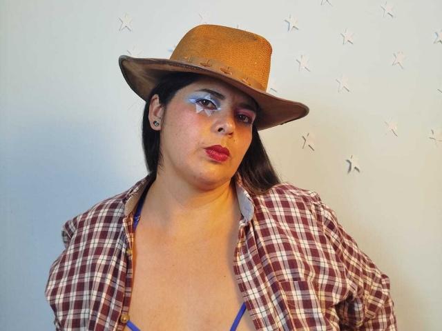 ZafiroYeux ha la pelle olivastra, occhi marroni, rossetto rosso, ombretto blu, un cappello marrone e una camicia a quadri sopra un reggiseno blu, mostrando un viso paffuto con un'espressione accigliata..