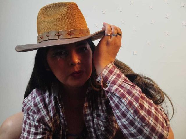 ZafiroYeux with long brown hair, wearing a tan straw hat with cross-stitched band, and a red plaid shirt, holding the hat with a serious, slightly pouty expression.