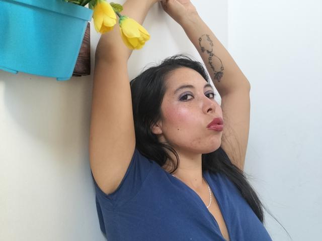 NikkieMilky with arms raised, pouting lips, dark hair, blue top, yellow roses in blue pot, tattoo on right arm, light skin, subtle makeup, slight blush.