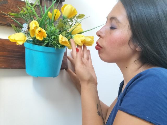 NikkieMilky with dark hair, blue shirt, and red lipstick, kisses a blue pot of yellow tulips, holding her hand up. She's got a tattoo on her left wrist.
