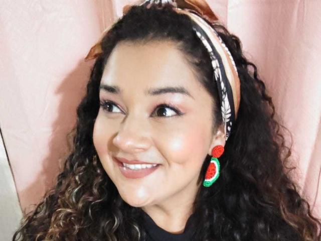NataliaRoa69 has curly dark hair, a brown headband, red and green earrings, and a smile showing her shiny skin and brown eyes. Her tits are not visible.