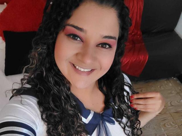 NataliaRoa69, curly black hair, smiling, pink eyeshadow, white and blue sailor top, red nails, holding shirt, looking at camera, white skin, cute, sexy, medium-sized tits.