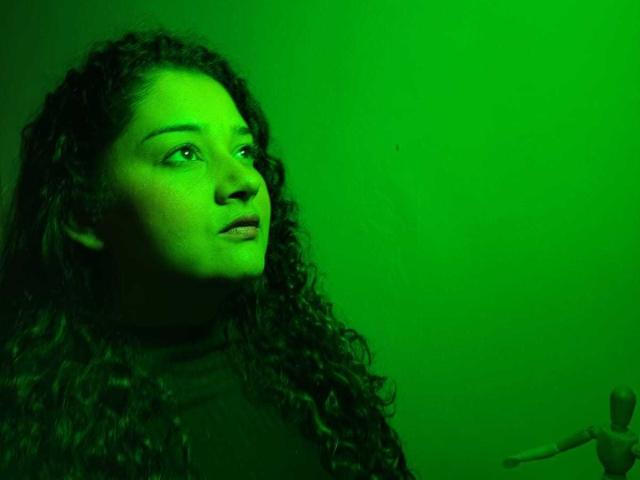 NataliaRoa69 with curly dark hair, green-tinted face, looking to the side, wearing a black top, holding a small object in her right hand.