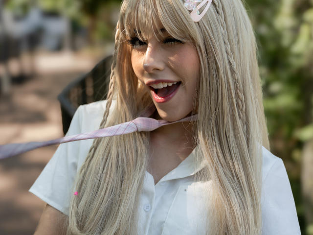 ValerieBunny with long blonde hair, white shirt, pink hair clip, and pink ribbon in mouth, smiling with pink lips, soft sunlight, and blurred background.