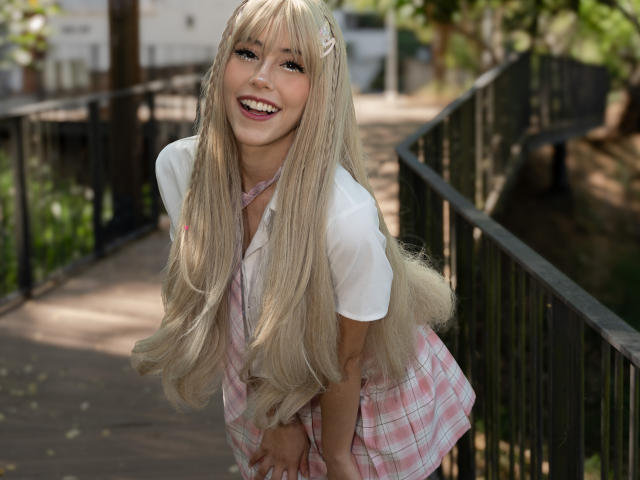ValerieBunny with long blonde hair, white shirt, pink plaid skirt, smiling, bending forward, showing off her tits, cute, outdoors, black metal railing, green trees in the background.