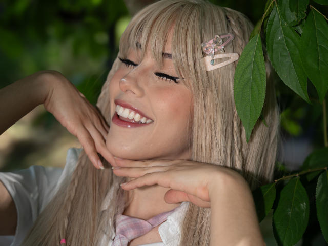 ValerieBunny with blonde hair, pink hairclip, white shirt, smiling, eyes closed, hands near chin, green leaves in background, soft natural light, cute, relaxed, happy expression, pretty.