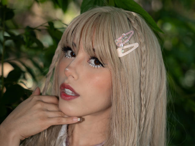 ValerieBunny has blonde hair with pink butterfly hairpins, brown eyes, and red lipstick. Her hand touches her cheek, looking up with a slightly open mouth.