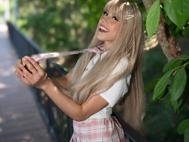 ValerieBunny with long blonde hair, white shirt, pink plaid skirt, holding pink tie, smiling, standing by tree, leaf in hair, hands near tits, green background.