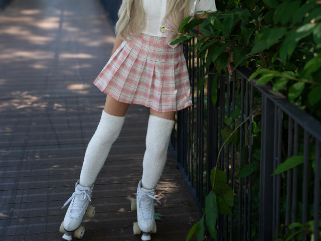 ValerieBunny's ass in a pink plaid skirt, white knee socks, and white roller skates, standing by a black fence with green leaves. Her blonde hair is visible.