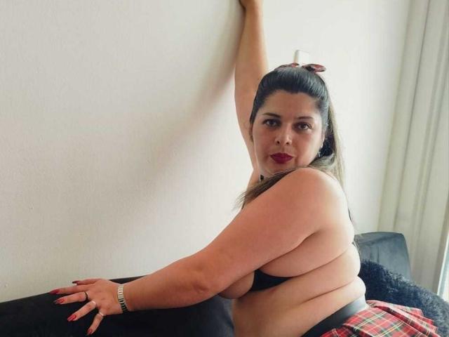 ZafiroYeux con grandi seni, braccio sollevato, rossetto rosso, reggiseno nero, gonna a quadri rossa, divano nero, unghie lunghe, braccialetto e capelli scuri raccolti in una coda di cavallo..