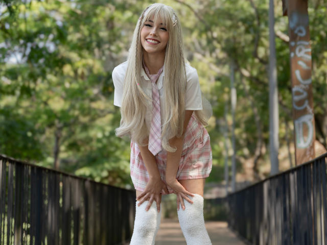 ValerieBunny with long blonde hair, smiling, wearing a pink plaid shirt, white thigh-high socks, and a pink necktie, bending forward, hands on thighs, in a park.