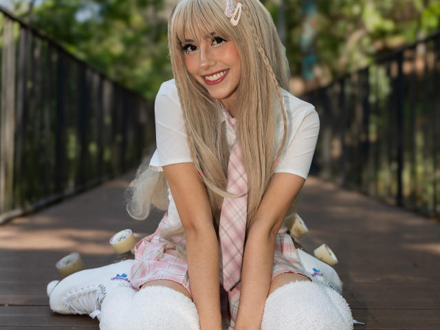  ValerieBunny with long blonde hair, smiling, wearing a pink plaid dress, white shirt, and white thigh-high socks, kneeling on a wooden path, surrounded by fluffy white toys.