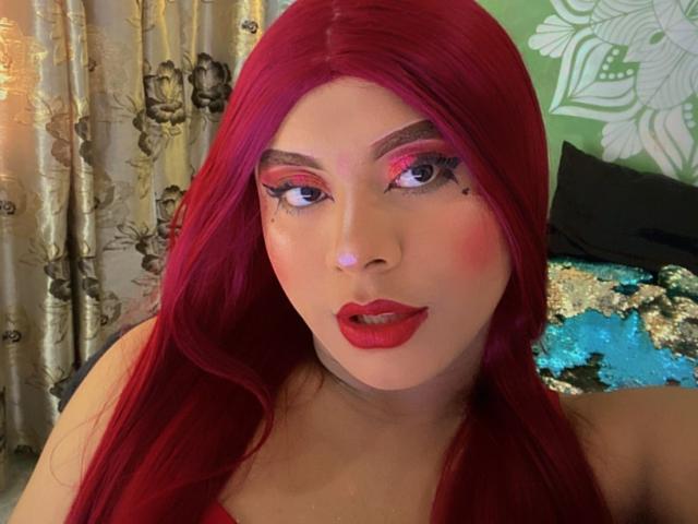 EmiliRoux with bright red hair, pink eye makeup, red lipstick, and a slightly open mouth. Her skin is fair, and she has small beauty marks on her right cheek.