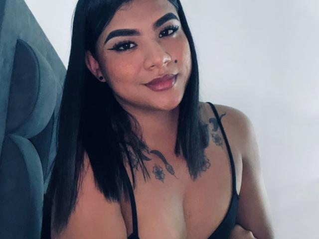 SammyJohnison has black hair, a cute smile, and small tits with tattoos above her chest. She's wearing a black spaghetti strap top and has black ear gauges.