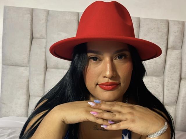 KorinaKova with long black hair, red hat, red lipstick, and painted nails, resting her hands on her chin, showing a tattoo under her neck.