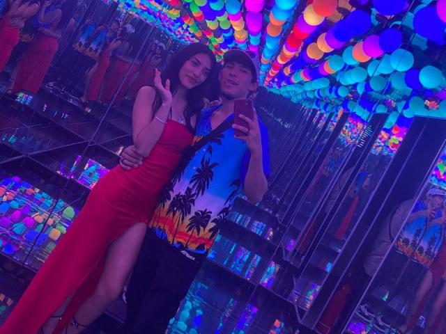HotSweetLovers in a red dress with a thigh slit, and HotSweetLovers in a blue shirt with palm trees, taking a selfie in a neon-lit club.