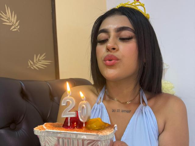 Daffne Addison, con i capelli castani, indossa una tiara dorata, un top a halter azzurro chiaro, labbra rosa carnose, tiene in mano una torta con candeline, con i numeri "20" e della frutta, una collana, un tatuaggio sul petto..