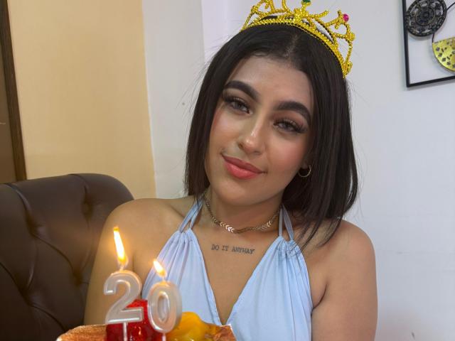 Daffne Addison, met zwart haar, draagt een gele kroon, een lichtblauwe haltertop en een gouden ketting, en glimlacht naar kaarsen met het nummer "20". Haar borsten zijn klein en haar achterkant is niet zichtbaar..
