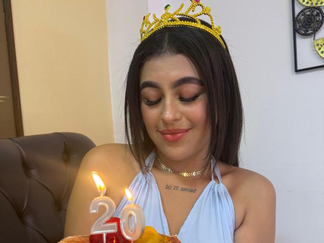 DaffneAddison with dark hair, wearing a gold tiara, blue halter top, and red lipstick, has lit candles numbered "20" in front of her, smiling softly.