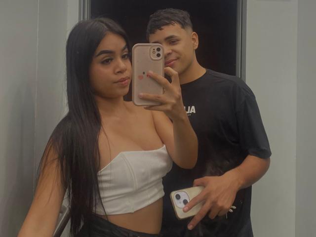 Isabella et Matteo, avec de longs cheveux noirs, Isabella porte un haut blanc moulant qui dévoile ses seins, un pantalon noir, et se tient à côté de Matteo qui porte un t-shirt noir. Elle tient un téléphone pour prendre un selfie devant un miroir..