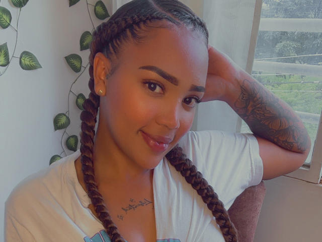 MorganaX with braided brown hair, tattoo on right arm, white shirt, smiling, hand in hair, earrings, soft light, window in background, green leaf vine on wall.
