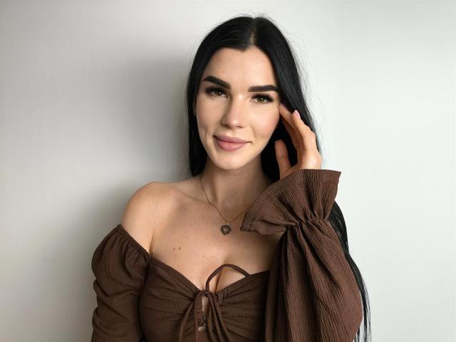 SofiaDream with long black hair, brown off-shoulder top, heart necklace, smiling, hand touching hair, subtle makeup, light skin, medium-sized tits, attractive, soft light, relaxed pose.