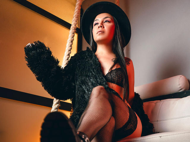 CharlotteJoliie with black hat, lace bra, fishnet stockings, and black fur over her tits, sitting on white couch, holding fluffy black toy, looking up with red makeup.