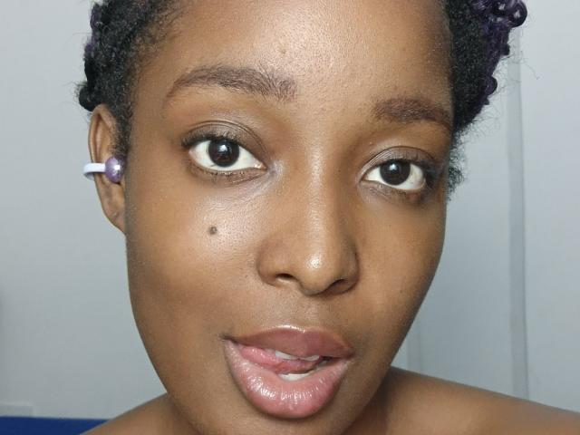 ChelseyeBonny's face with dark brown skin, large eyes, purple hair buns, white ear clip, slight smile, glossy pink lips, small beauty mark on her left cheek.