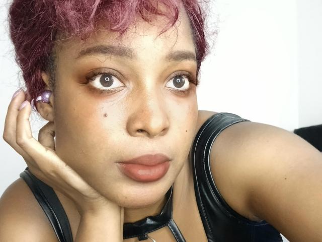 ChelseyeBonny with pink curly hair, brown eyes, and full lips, wearing a black leather top, resting her head on her hand, showing a small mole on her right cheek.