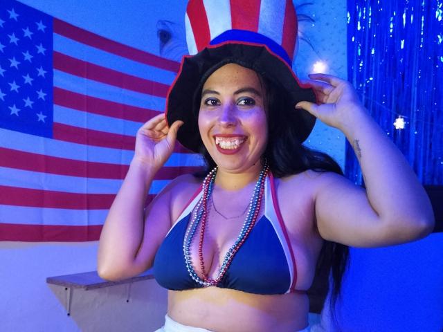 NikkieMilky with dark hair, wearing a star-spangled hat, blue bikini top, and bead necklace, smiling, holding hat, showing her tits, and standing with a playful expression.