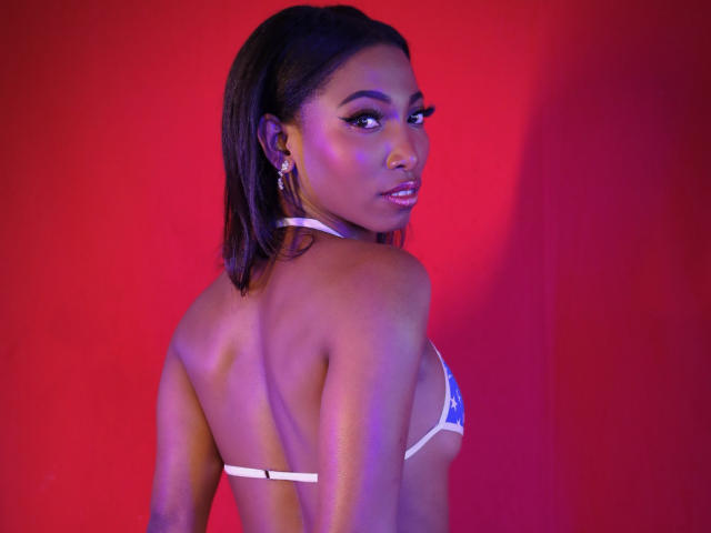 SabrinaDevil with dark skin, shiny brown hair, wearing a tiny white bikini, showing her back and looking over her shoulder with a sultry expression. Her tits are small.