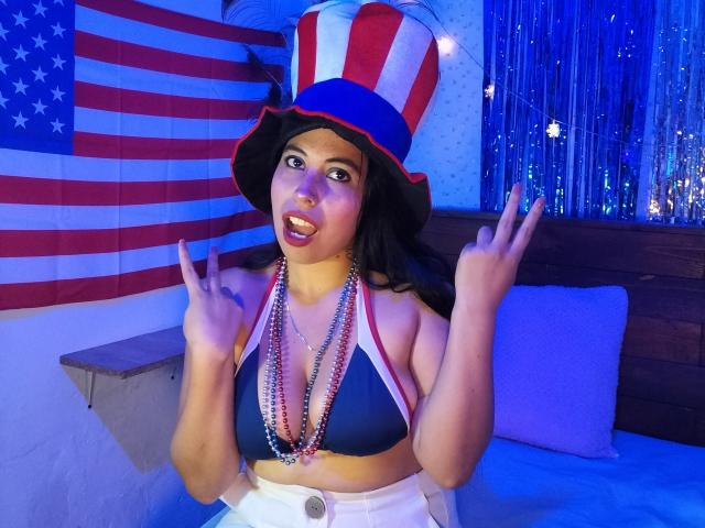 NikkieMilky with dark hair, wearing a red-white-blue top hat, blue bikini top, white pants, bead necklace, making double peace signs, blue-tinted room, American flag in background.