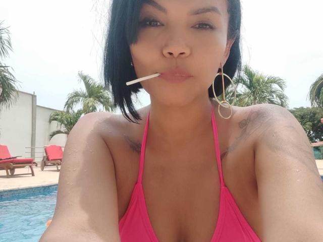 ExoticLyla mit schwarzen Haaren, einem pinken Bikini-Oberteil und einer Zigarette im Mund, die ihre Brüste und Tattoos zeigt, mit großen Reifenohrringen, am Pool..