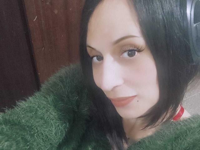 CharlottePrettyHot has pale skin, black hair, and is wearing green fuzzy clothing. She's looking at the camera with a slight smile, her long eyelashes are prominent.