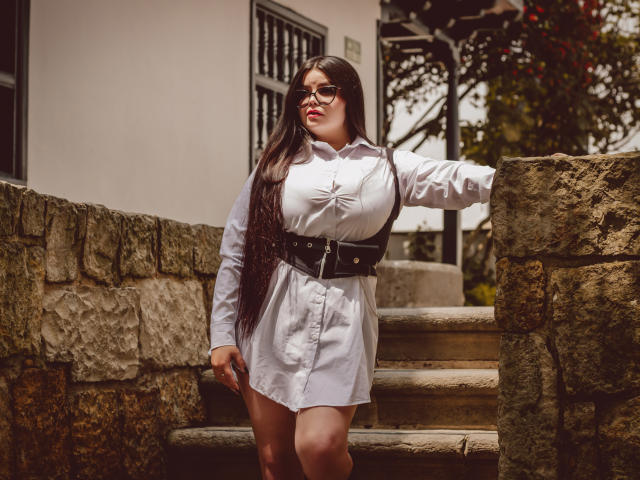 BrithneyLee with long black hair, glasses, white button-up dress, black belt, big tits, standing on stone steps, hand on wall, sunlight, looking to the side, confident.