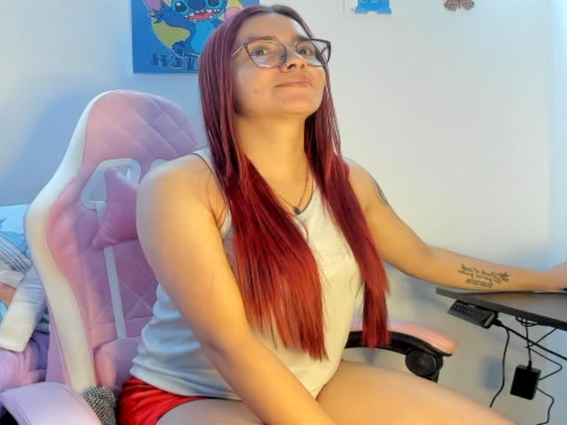 EimmyHoskX with red hair, glasses, white tank top, red shorts, sitting in pink gaming chair, tattoo on right forearm, looking up with a slight smile, hands on desk.