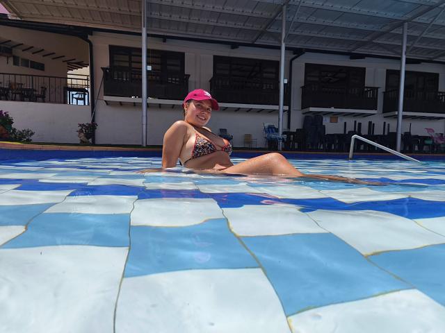 ZoeTobon69, with a pink cap, is in a blue and white tiled pool, wearing a leopard print bikini, showing her tits and ass, smiling, with water up to her thighs.